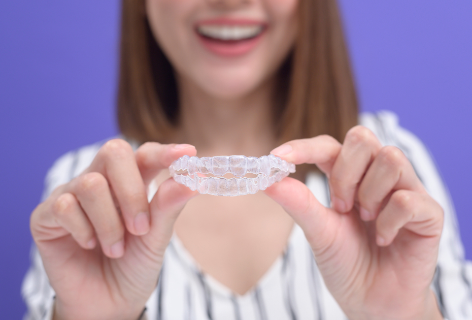 Woman smiling and holding clear aligners in her hands against a purple background, showcasing invisible teeth straightening treatment by Smileie.