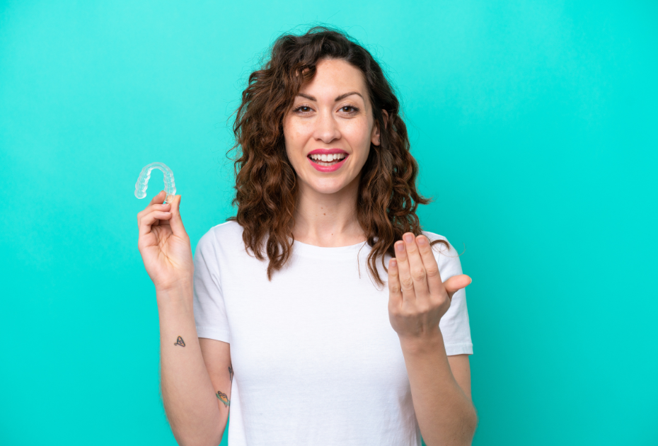 Smiling woman holding clear aligners in hand on turquoise background, showing her teeth aligner during Smileie treatment.