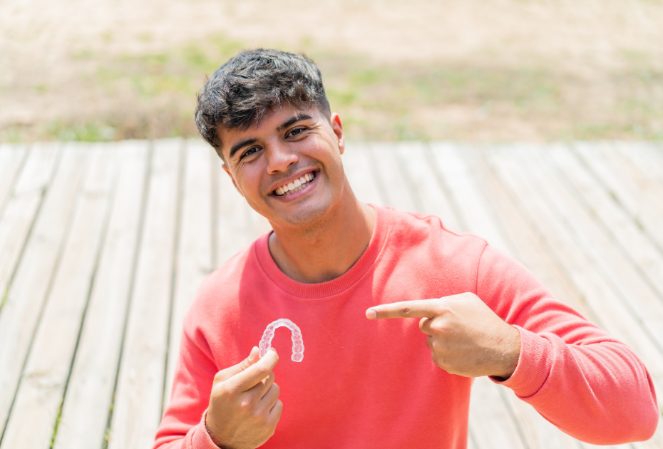 Smiling young man holding a clear aligner tray and pointing to it outdoors.