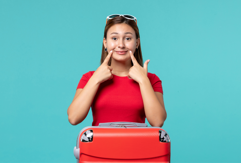 Smiling young woman with suitcase pointing at her teeth, representing international travel with clear aligners.