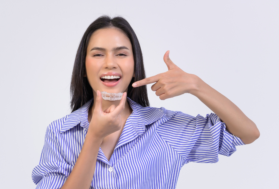 Woman smiling and holding clear aligners, pointing at her teeth while wearing invisible braces.