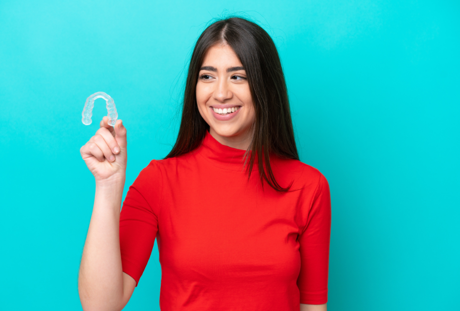 Smiling young woman holding a clear dental aligner against a teal background, representing Smileie clear aligners for comfortable at-home teeth straightening.